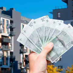 Dłoń trzyma banknoty 100 zł na tle nowoczesnych bloków mieszkalnych. Zastanawiasz się, jakie koszty można odliczyć przy sprzedaży mieszkania?