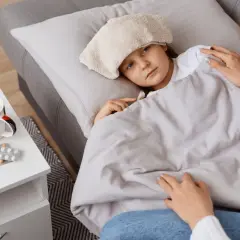 Chora dziewczynka z okładem na głowie leży pod kołdrą. Obok leki i kubek. To obraz troskliwej opieki na członka rodziny.