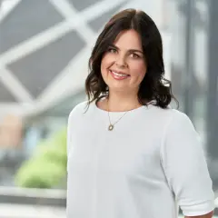 Uśmiechnięta kobieta w białej bluzce, z wisiorkiem w kształcie słońca, wspiera fundację rodzinną.