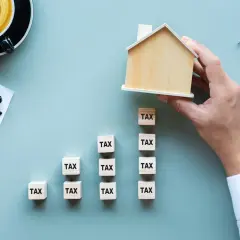 Działka budowlana niezabudowana. Klocki z napisem "TAX" tworzą wykres wzrostu, a ręka kładzie na szczycie drewniany domek.