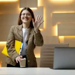 Uśmiechnięta kobieta w okularach macha ręką, trzymając teczkę i kawę. Czy wspólnik może pracować dla spółki bez umowy?