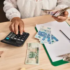 Kobieta liczy pieniądze na kalkulatorze, porównując dochody z samozatrudnienia a umowa o pracę. Na stole polskie banknoty, terminal płatniczy i dokumenty.