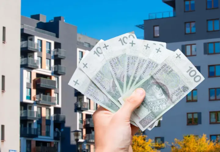 Dłoń trzyma banknoty 100 zł na tle nowoczesnych bloków mieszkalnych. Zastanawiasz się, jakie koszty można odliczyć przy sprzedaży mieszkania?