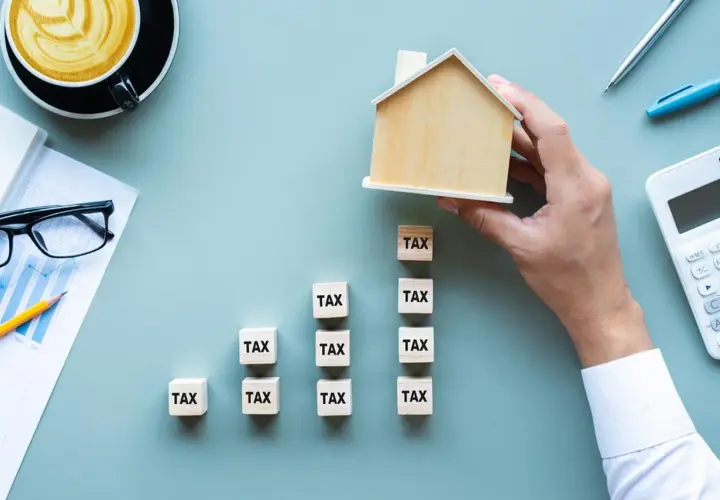 Działka budowlana niezabudowana. Klocki z napisem "TAX" tworzą wykres wzrostu, a ręka kładzie na szczycie drewniany domek.