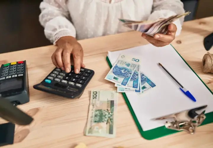 Kobieta liczy pieniądze na kalkulatorze, porównując dochody z samozatrudnienia a umowa o pracę. Na stole polskie banknoty, terminal płatniczy i dokumenty.