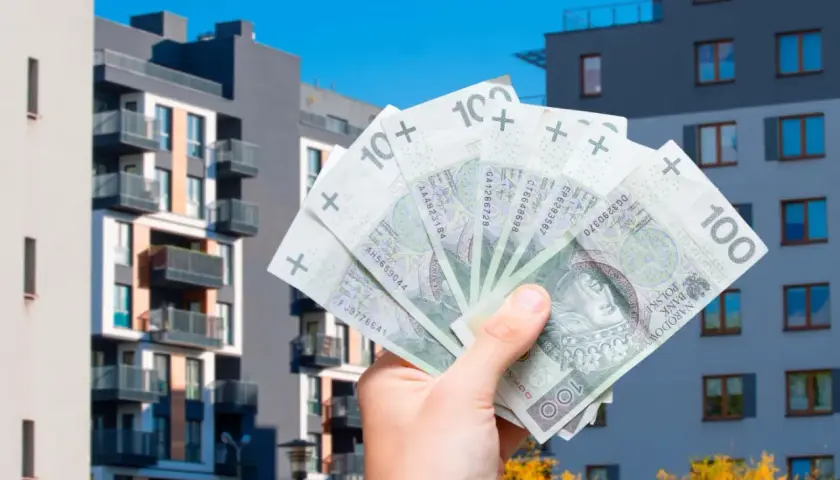 Dłoń trzyma banknoty 100 zł na tle nowoczesnych bloków mieszkalnych. Zastanawiasz się, jakie koszty można odliczyć przy sprzedaży mieszkania?