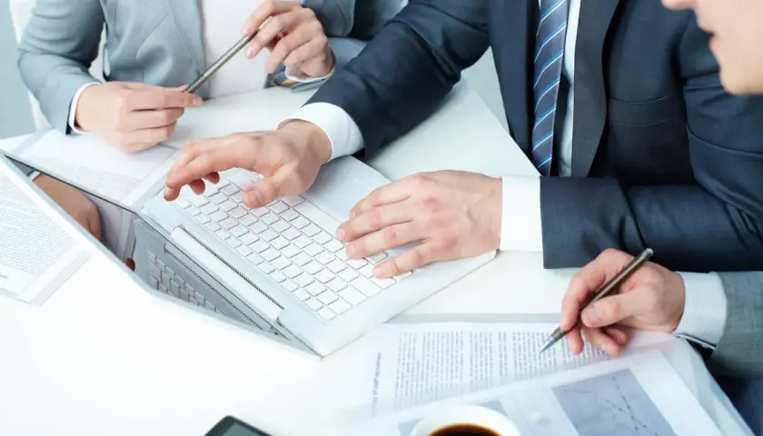 Biznesmeni analizują dokumenty, dyskutując o wynagrodzeniu prokurenta. Na stole laptop, kawa i telefon.