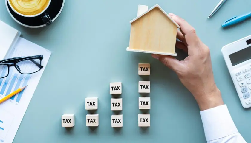 Działka budowlana niezabudowana. Klocki z napisem "TAX" tworzą wykres wzrostu, a ręka kładzie na szczycie drewniany domek.