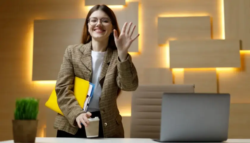 Uśmiechnięta kobieta w okularach macha ręką, trzymając teczkę i kawę. Czy wspólnik może pracować dla spółki bez umowy?