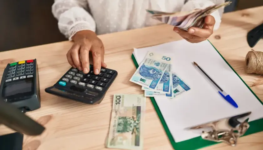 Kobieta liczy pieniądze na kalkulatorze, porównując dochody z samozatrudnienia a umowa o pracę. Na stole polskie banknoty, terminal płatniczy i dokumenty.