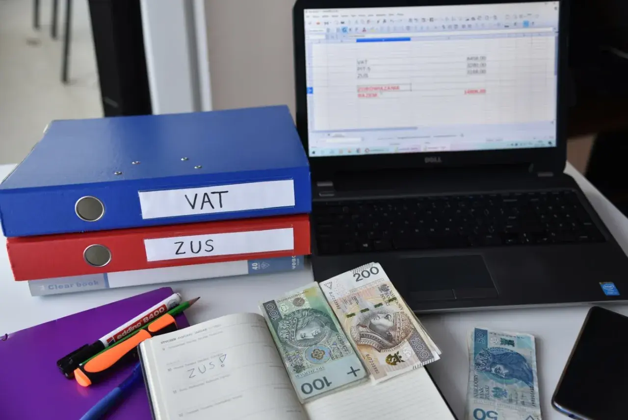 Segregator VAT i ZUS, laptop z arkuszem kalkulacyjnym, pieniądze i notatnik. To obrazuje, co to znaczy być vatowcem na ryczałcie.