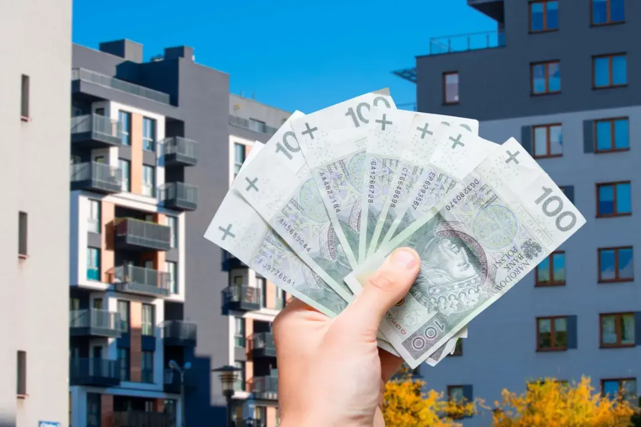 Dłoń trzyma banknoty 100 zł na tle nowoczesnych bloków mieszkalnych. Zastanawiasz się, jakie koszty można odliczyć przy sprzedaży mieszkania?