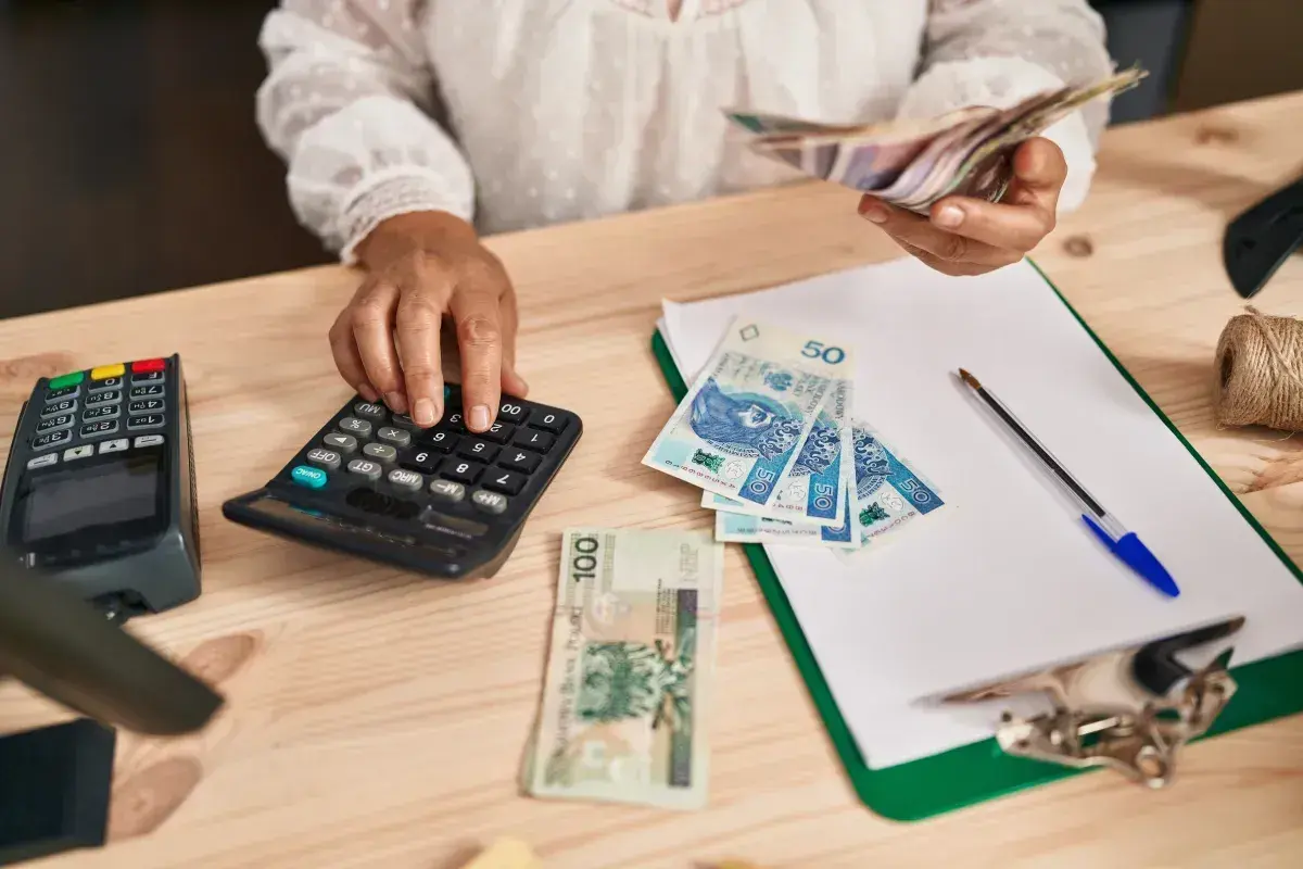 Kobieta liczy pieniądze na kalkulatorze, porównując dochody z samozatrudnienia a umowa o pracę. Na stole polskie banknoty, terminal płatniczy i dokumenty.