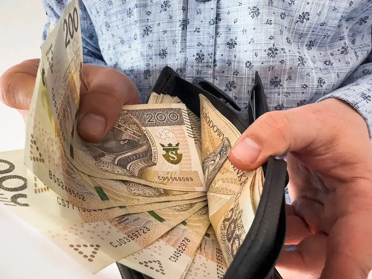 Dłonie trzymają portfel z banknotami 200 zł. Może to być nagroda jubileuszowa za wieloletnią pracę.